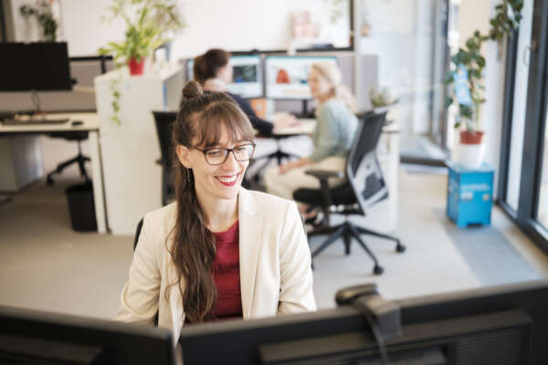 Eine Frau sitzt an einem Computer im Büro und lächelt.
