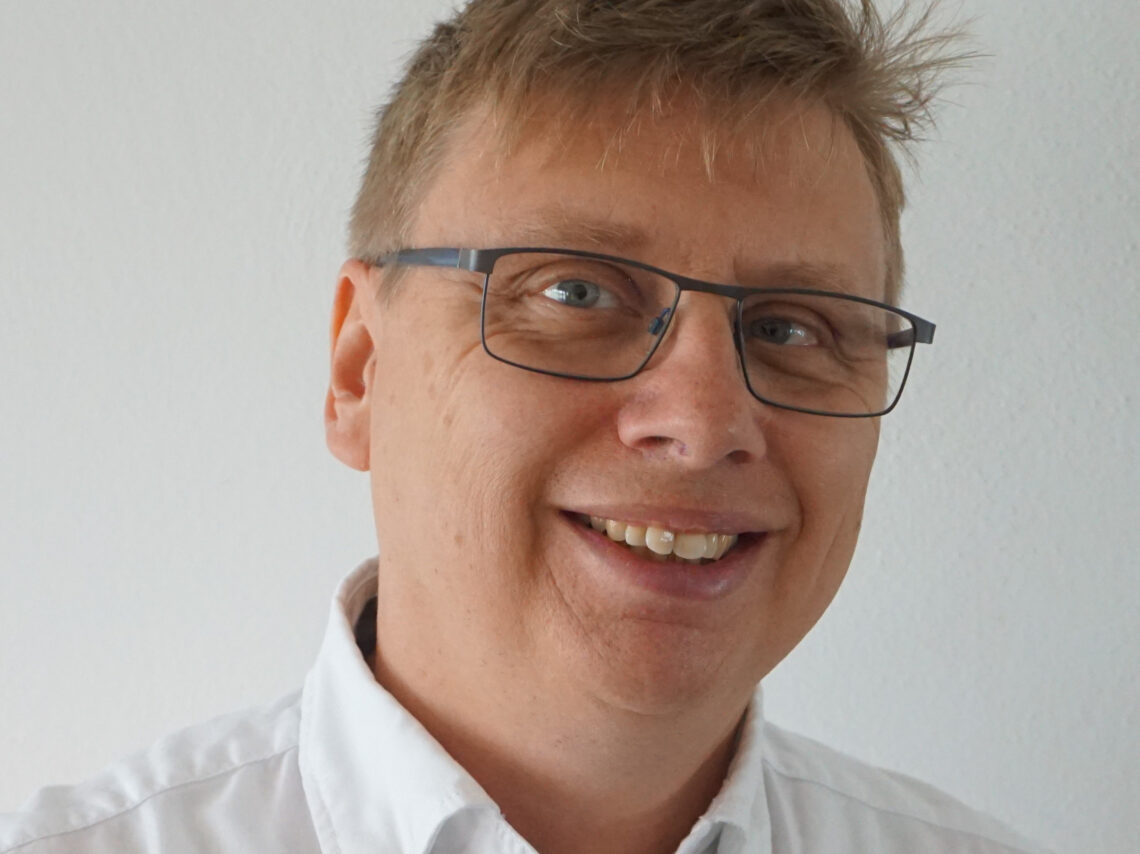 Lachendes Porträt von Roland Sigrist vor einer weißen Wand.