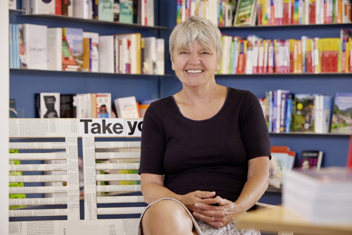 Sabine Gartmann sitzt in der Buchhandlung Die Schatulle vor einem Bücherregal und lächelt.