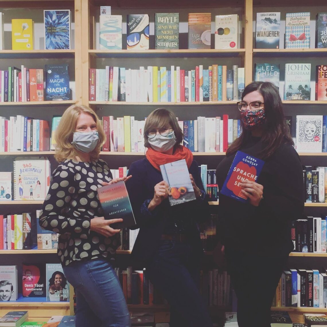 Das Team von Grimms Buchhandlung steht mit Buch in der Hand und Mundschutz vor einem Bücherregal.
