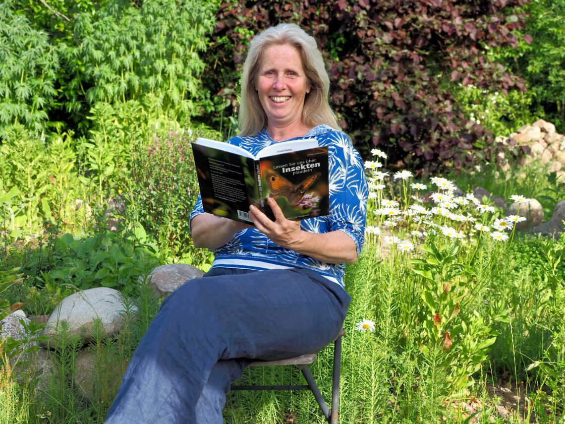 Ulrike Lohmann sitzt mit einem geöffnetem Buch in der Hand auf einem Stuhl im Garten umgeben von Grün.