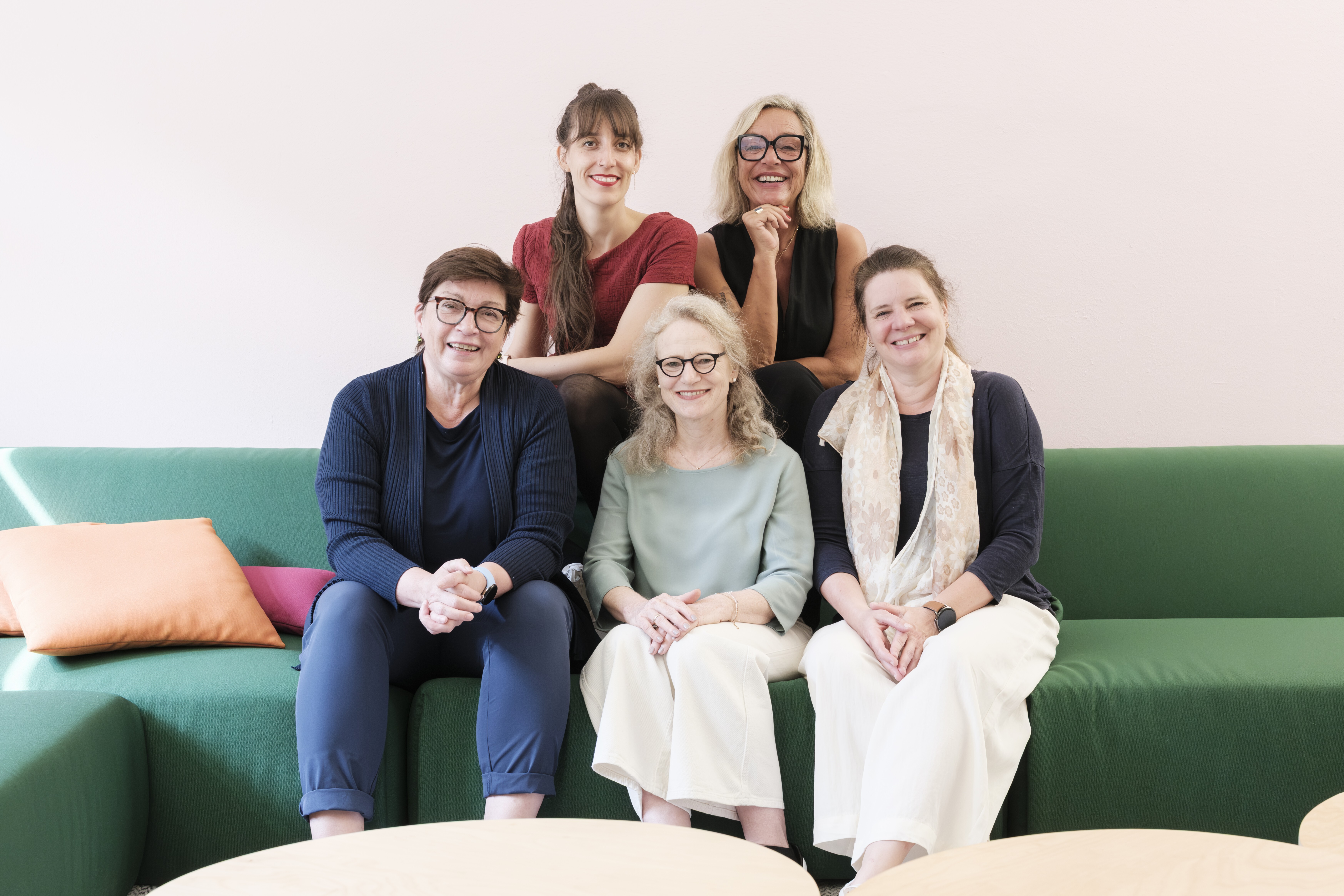 Fünf Frauen sitzen auf einer Couch und lächeln in die Kamera.