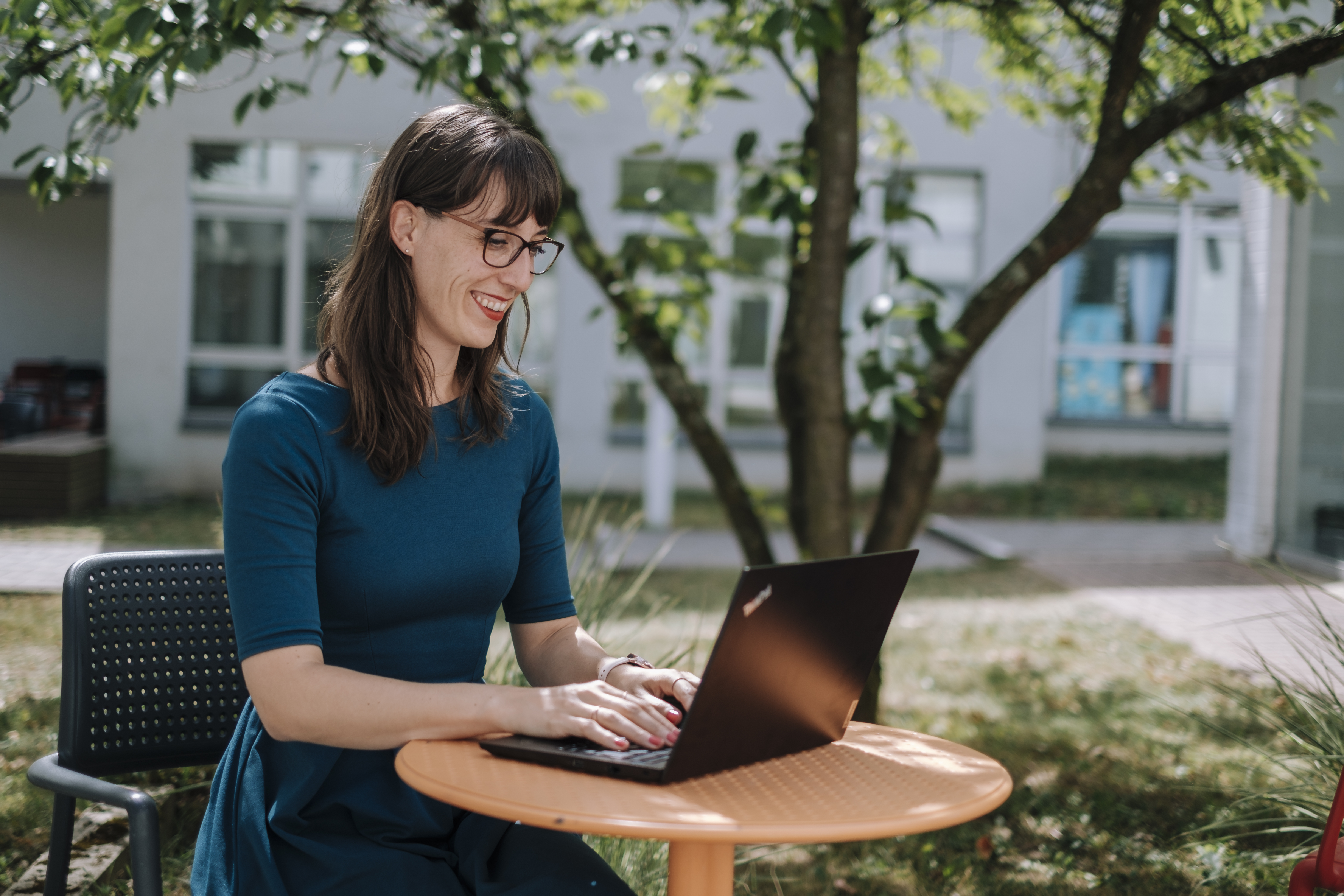 Rahel Trotta an einem Tisch im Freien. Sie sitzt lächelnd an ihrem Laptop.
