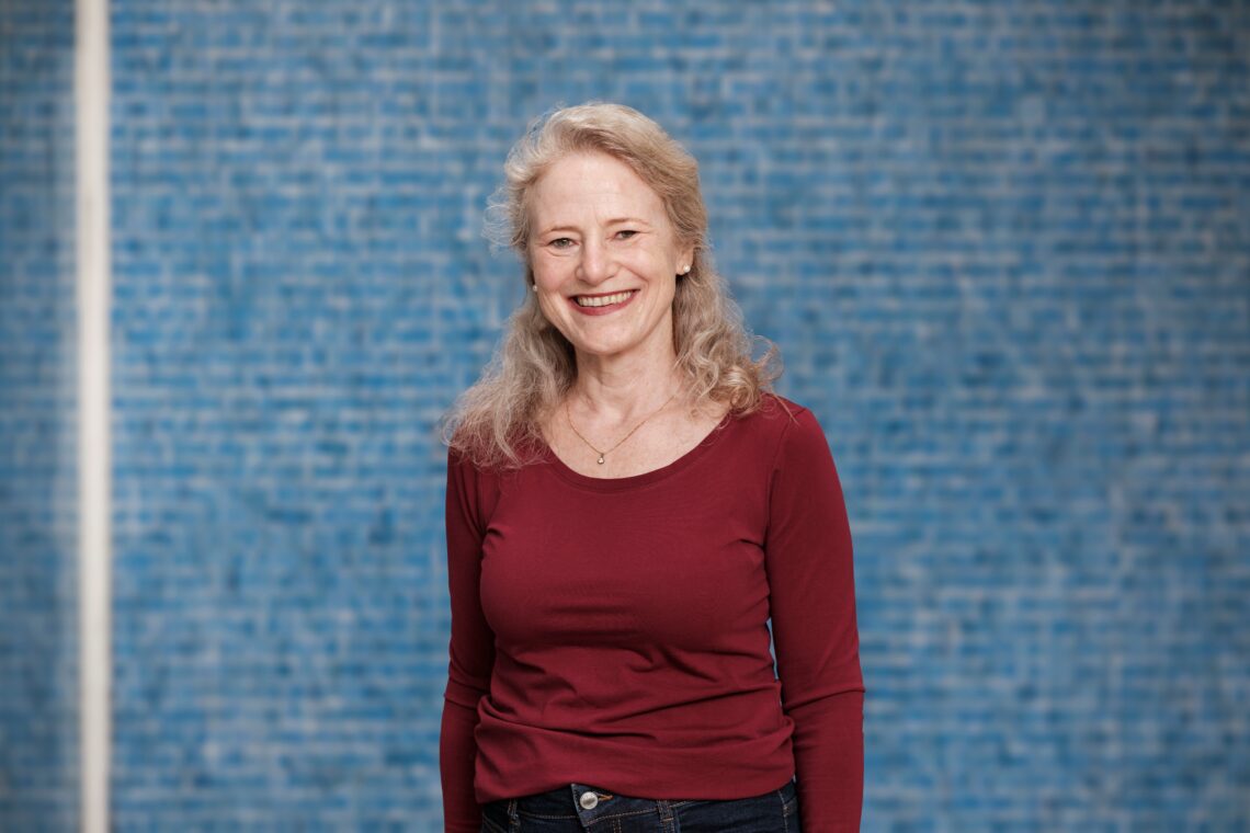 Lachendes Porträt von Susanne Fuchs vor einer dunkelblauen Mosaikwand.