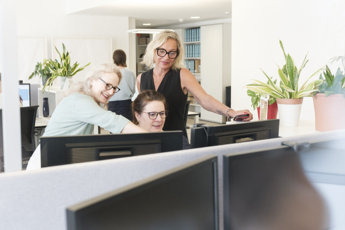 Zwei Frauen stehen neben einer sitzenden Frau an einem Schreibtisch und zeigen auf etwas auf dem Bildschirm.