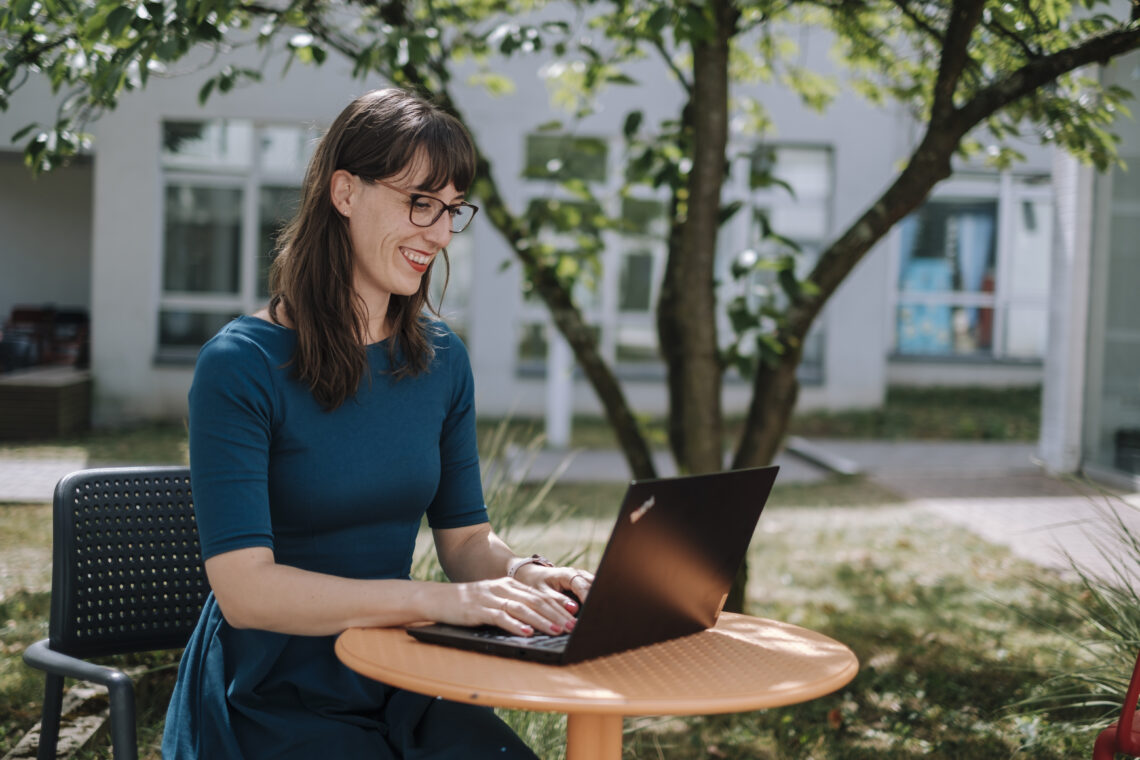 Eine Frau sitzt druaßen vor einem Baum an einem kleinen runden Tisch und arbeitet an einem Laptop.