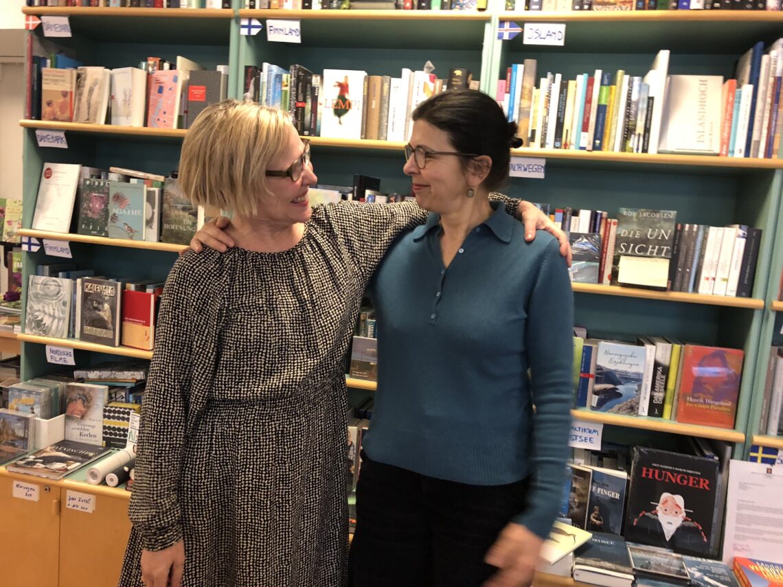 Zwei Frauen stehen in einer Buchhandlung vor einem Bücherregal und lächeln sich an.