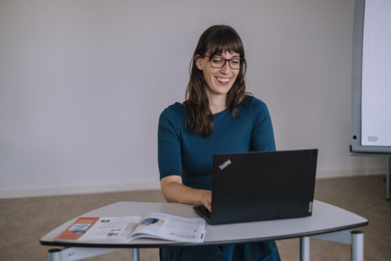 Eine Frau sitzt in einem Arbeitsraum an einem Tisch vor einem offenem Laptop.