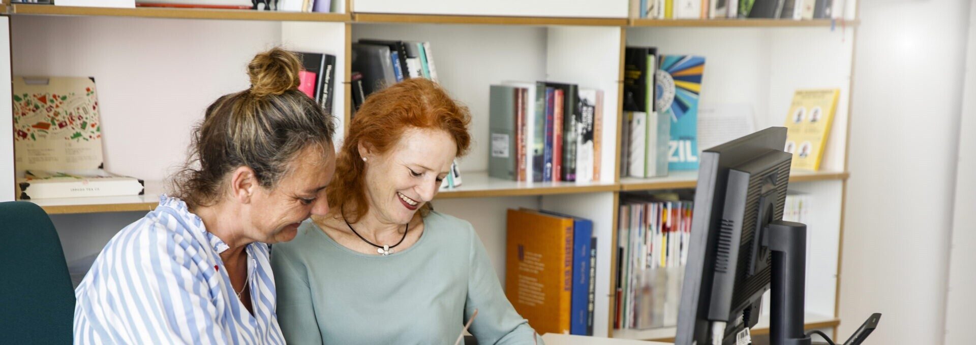 Zwei Frauen sitzen zusammen vor einem Regal an einem Schreibtisch vor einem Computer-Bildschirm.