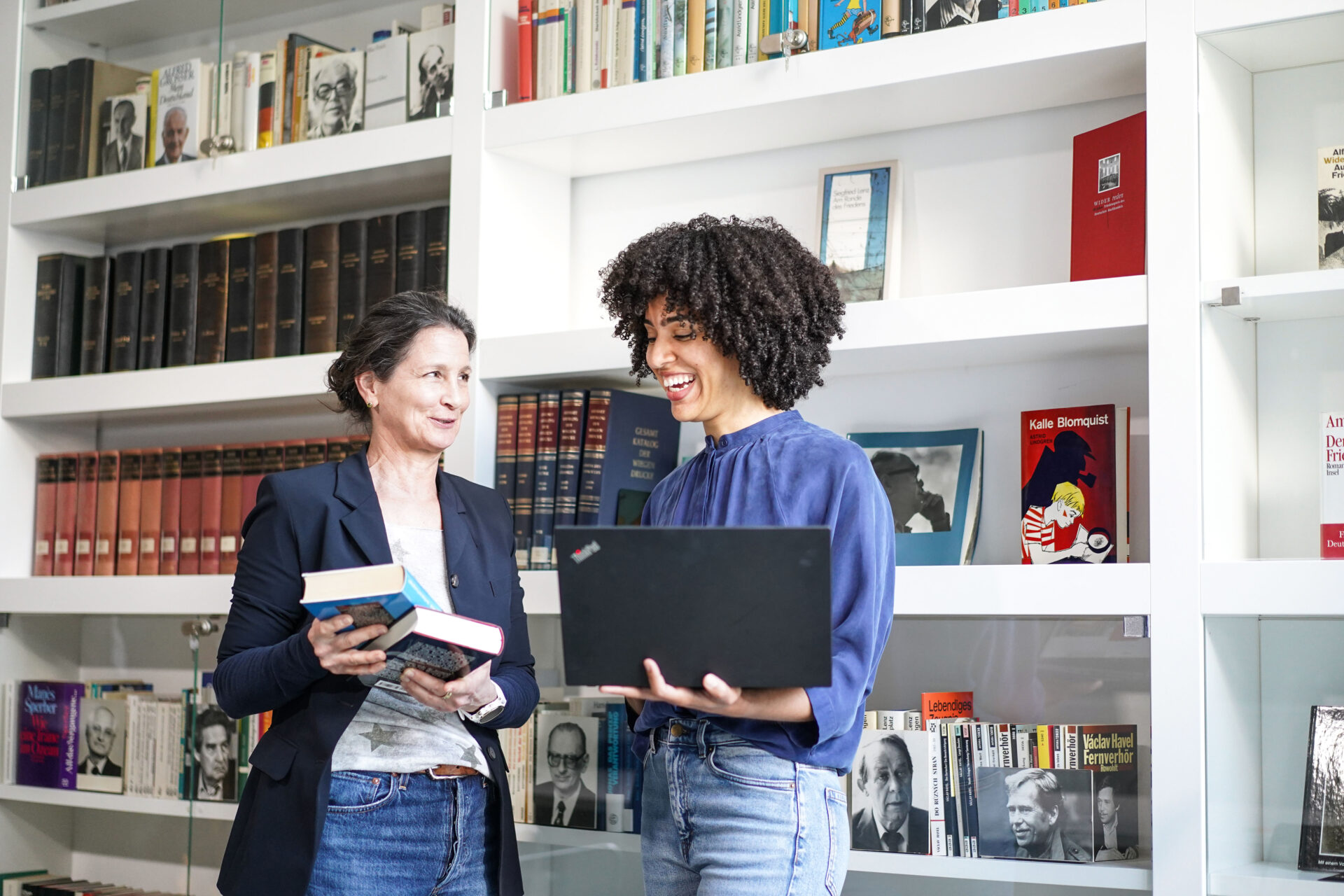 Zwei Frauen stehen vor einem Bücherregal während die linke Frau Bücher und die rechte Frau einen offenen Laptop hält.