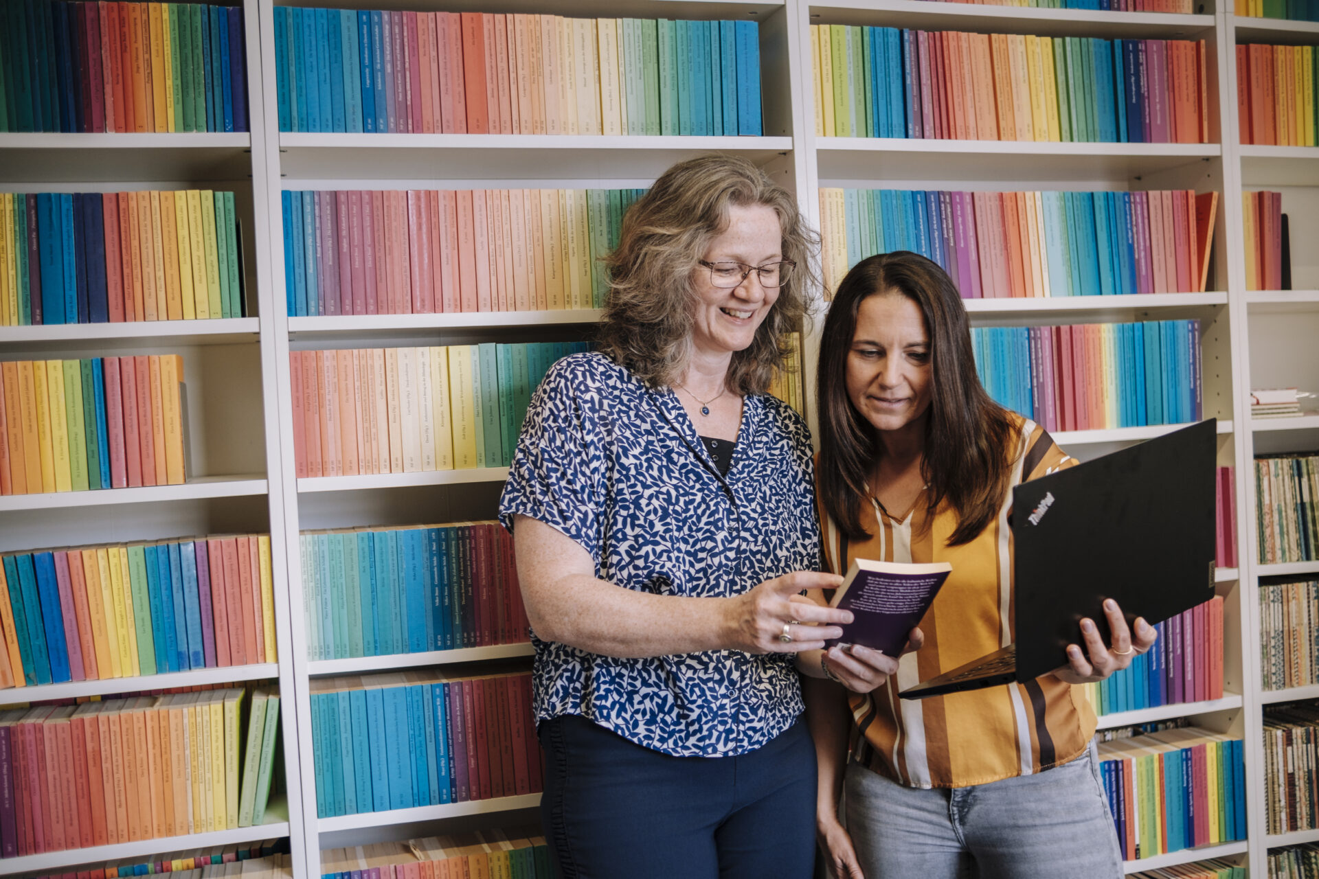 Zwei Frauen stehen vor einer Regenbogen-Bücherwand und schauen gemeinsam auf ein Buch und einen offenen Laptop.
