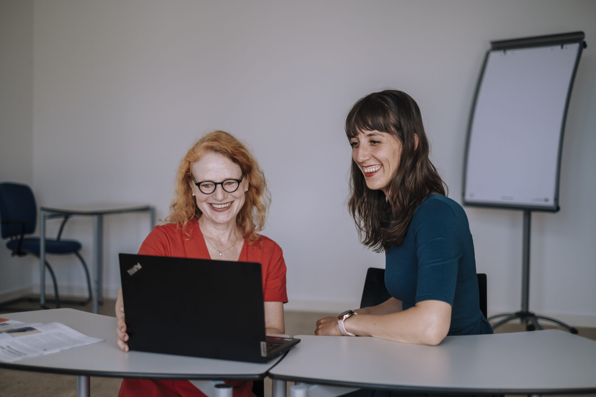 Zwei Frauen sitzen in einem Arbeitsraum an einem Tisch vor einem offenem Laptop.