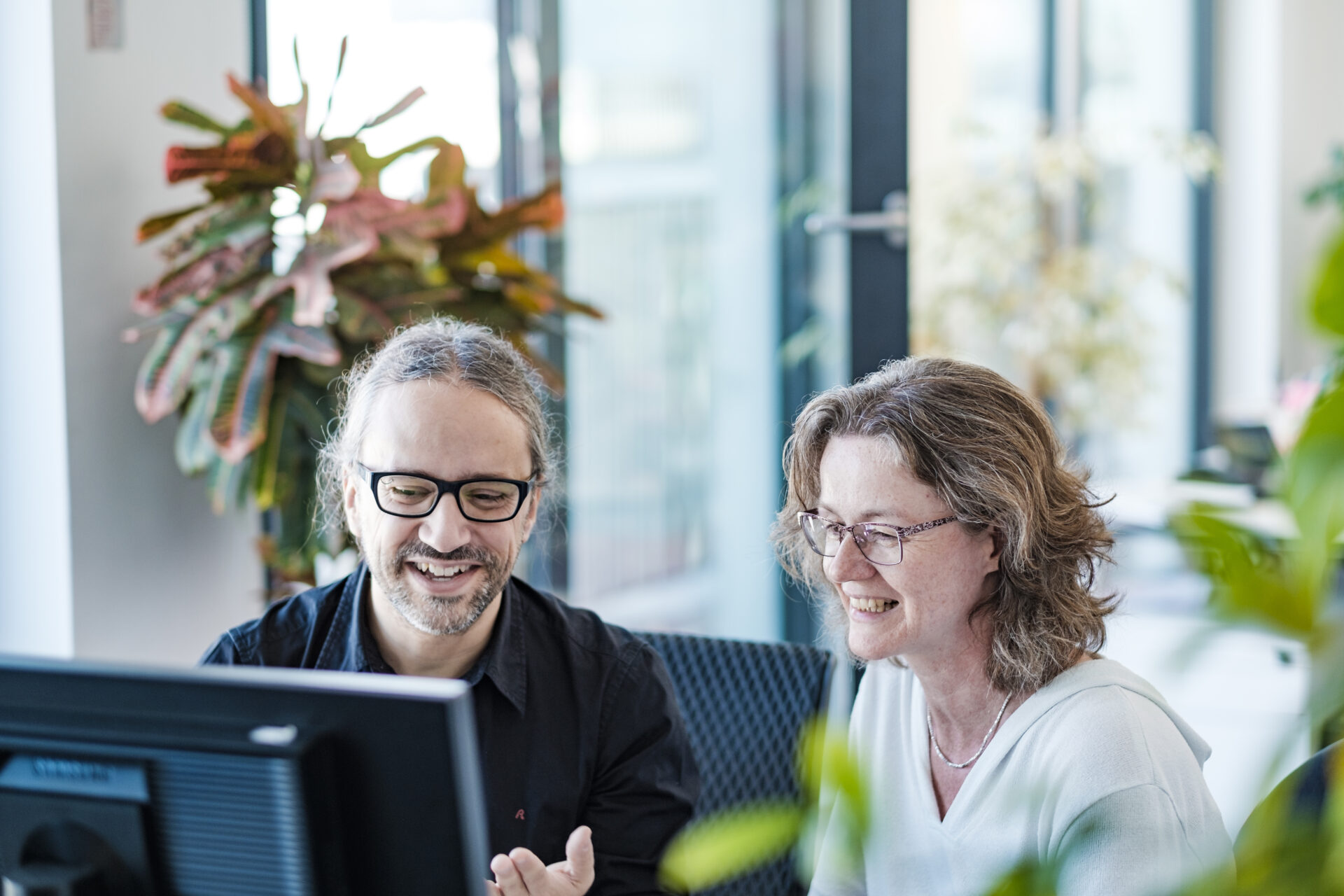 Ein Mann und eine Frau sitzen zusammen an einem Schreibtisch vor einem Computer, im Hintergrund sind Pflanzen.