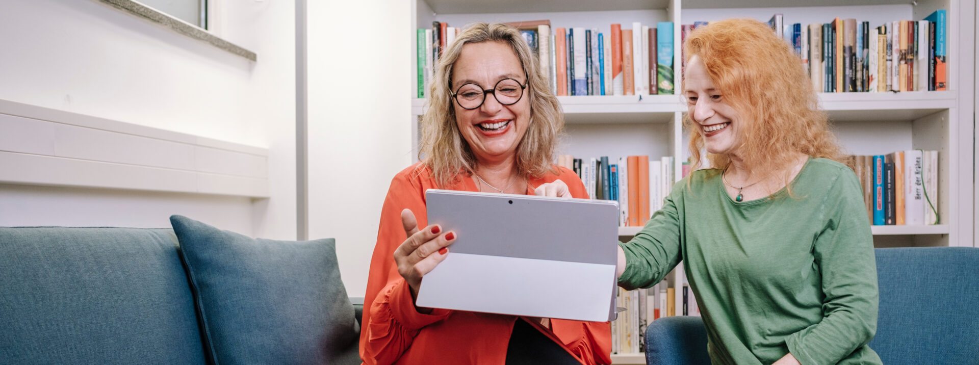 Zwei Frauen sitzen auf einer Couch vor einem Bücherregal und die linke Frau hat einen Laptop auf ihrem Schoß.