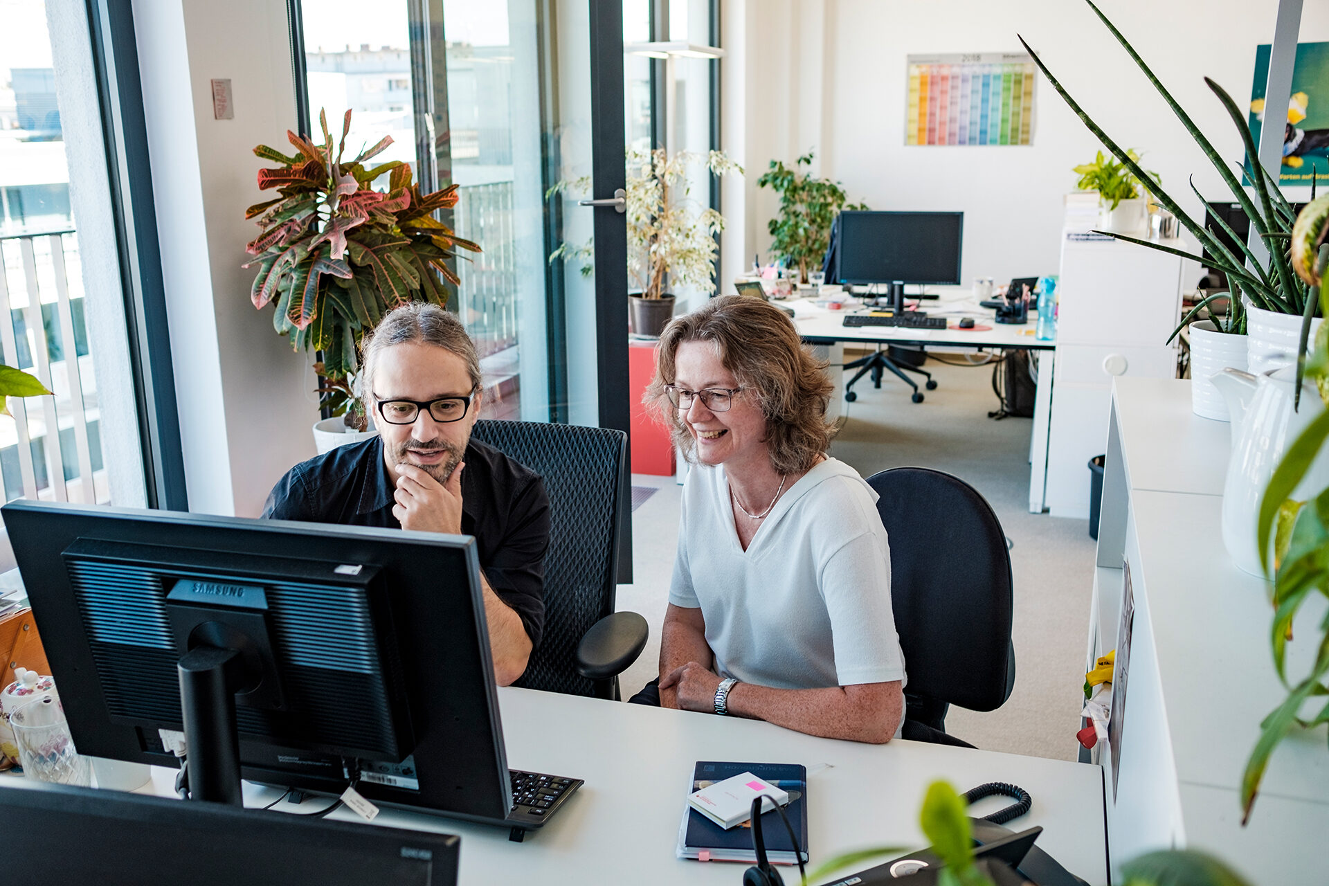 Ein Mann und eine Frau sitzen zusammen an einem Schreibtisch vor einem Computer-Bildschirm, im Hintergrund sind Pflanzen.