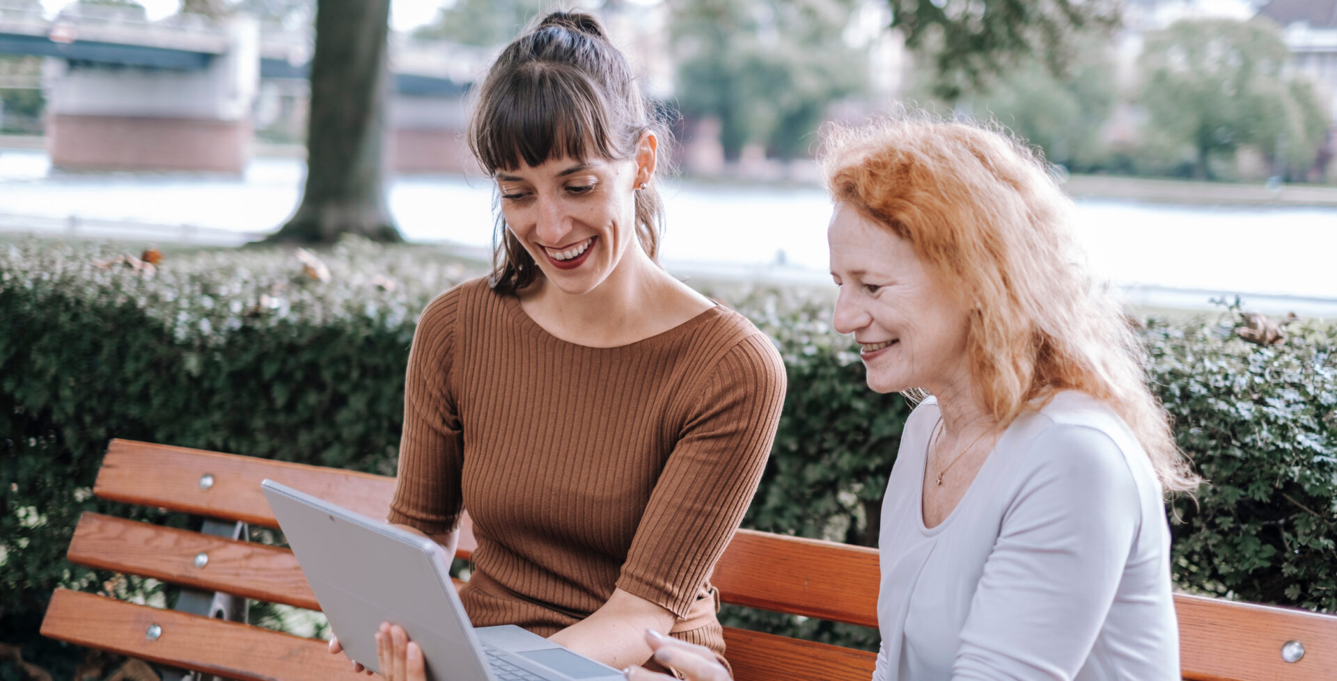 Zwei Frauen sitzen auf einer Bank am Main und tauschen sich vor einem offenen Laptop aus.