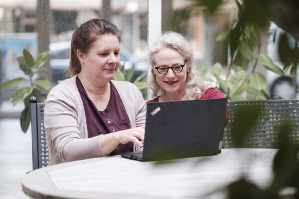 Zwei Frauen sitzen an einem Tisch vor einem Laptop und arbeiten daran.