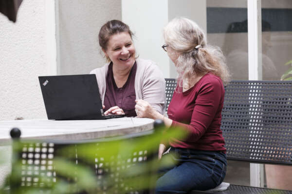 Zwei Frauen sitzen an einem Tisch vor einem Laptop und lächeln sich an.