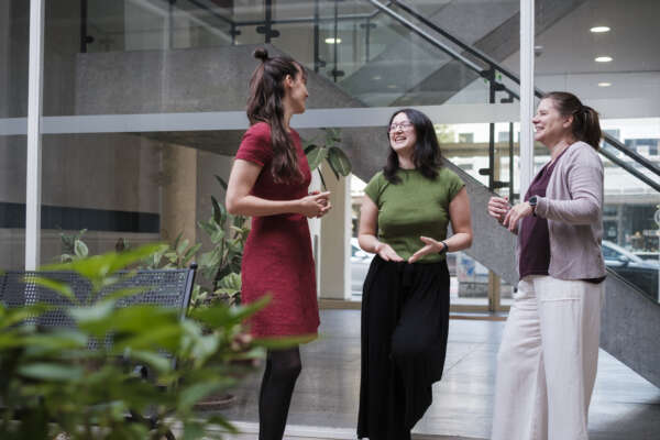 Drei Frauen stehen vor einer Glaswand in einem Innenhof und unterhalten sich.