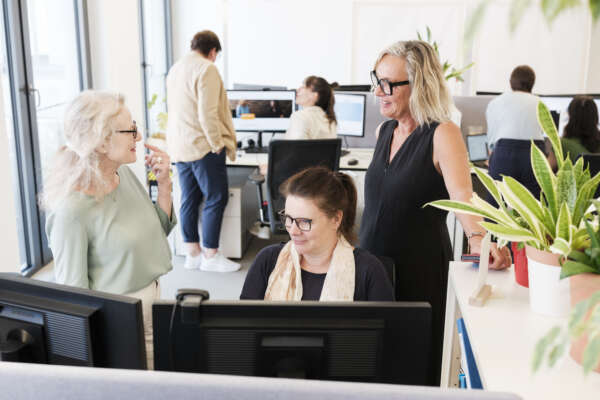 Zwei Frauen stehen hinter einer sitzenden Frau an einem Schreibtisch und unterhalten sich.