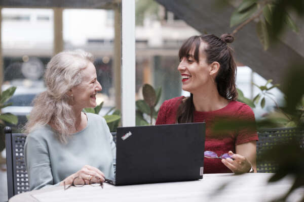 Zwei Frauen sitzen an einem Tisch vor einem Laptop und lächeln sich an.