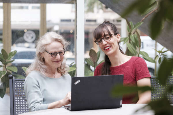 Zwei Frauen sitzen lächelnd an einem Tisch vor einem Laptop und arbeiten daran.