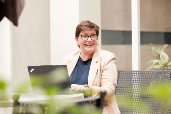 Eine Frau sitzt lachend vor einem Laptop an einem Tisch in einem Innenhof.