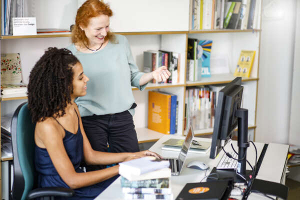 Eine Frau sitzt an einem Schreibtisch vor einem Bücherregal und links neben ihr steht eine weitere die mit ihr im Austausch ist.