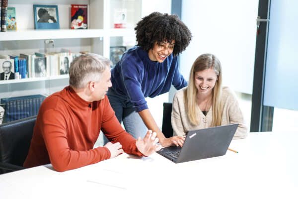 Ein Mann und eine Frau sitzen an einem Tisch vor einem offenen Laptop und zwischen ihnen steht eine weitere Frau die den Laptop bedient.