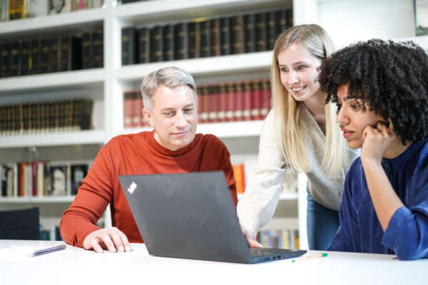 Ein Mann und eine Frau sitzen an einem Tisch vor einem offenen Laptop und zwischen ihnen steht eine weitere Frau die den Laptop bedient.