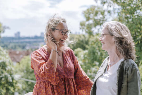 Zwei Frauen stehen auf einem Balkon und lächeln sich an, im Hintergrund sind Bäume.