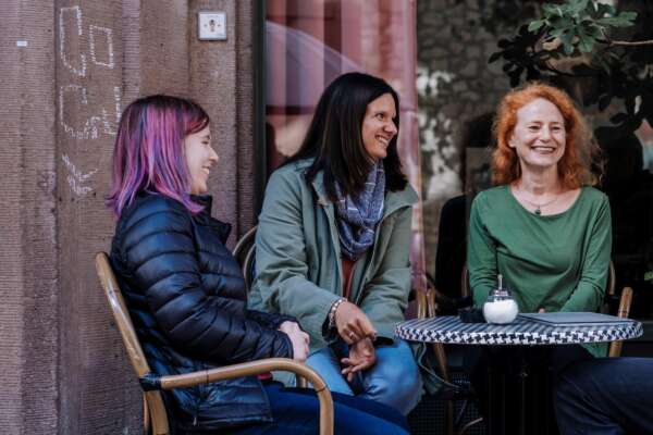 Drei lachende Frauen sitzen an einem runden Tisch eines Cafés.