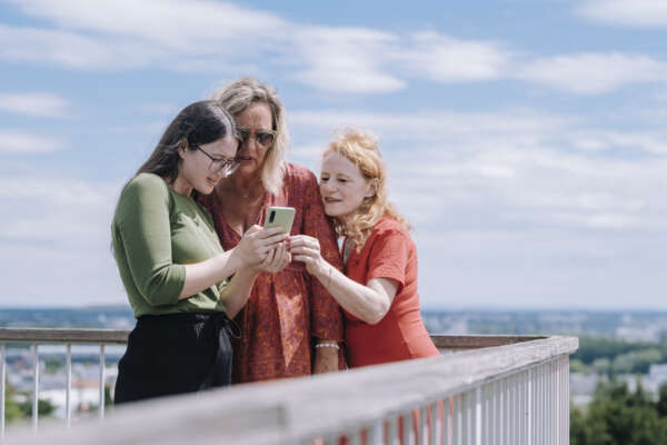 drei Frauen stehen in der Ecke eines Balkons und schauen zusammen auf ein Handy.