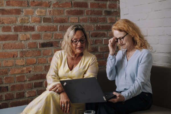 Zwei Frauen sitzen auf einem Sofa und schauen auf einen offenen Laptop der auf den Knien der rechten Frau liegt.