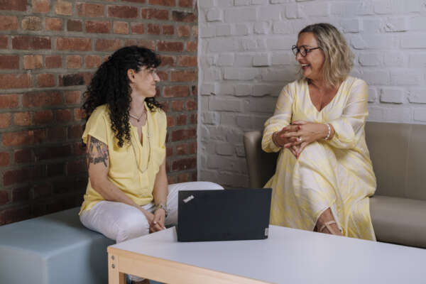 Zwei Frauen sitzen in einer Ecke auf Sofa-Möbeln und haben vor sich auf einem Couchtisch einen offenen Laptop stehen.
