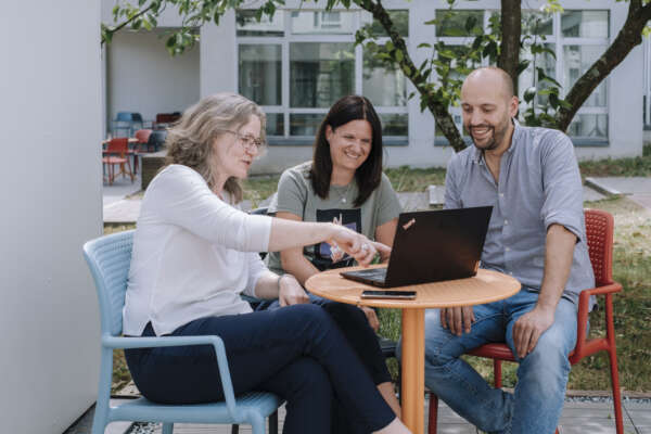 Zwei Frauen und ein Mann sitzen draußen vor einem offenem Laptop an einem kleinen runden Tisch.