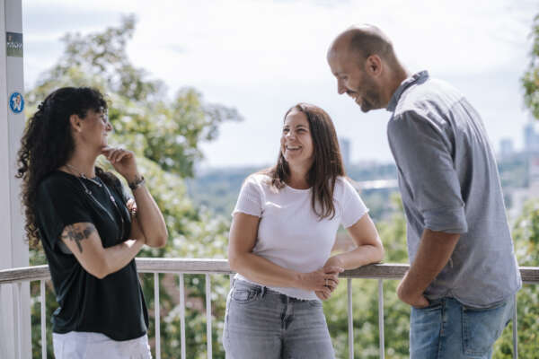 Zwei Frauen und ein Mann stehen an dem Geländer eines Balkons und tauschen sich aus.