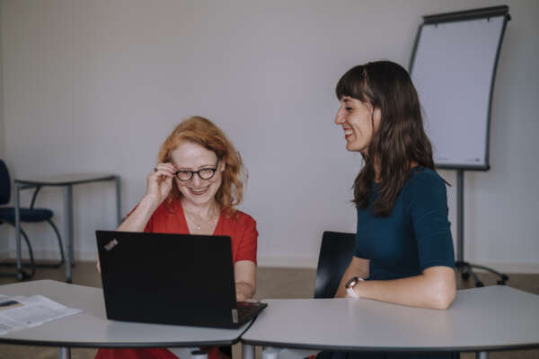Zwei Frauen sitzen in einem Arbeitsraum an einem Tisch vor einem offenem Laptop.