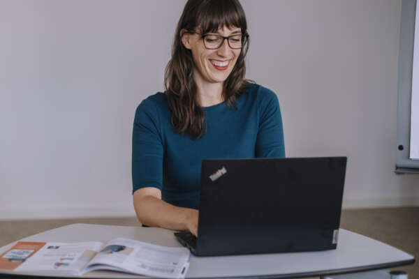 Eine Frau sitzt in einem Arbeitsraum an einem Tisch vor einem offenem Laptop.