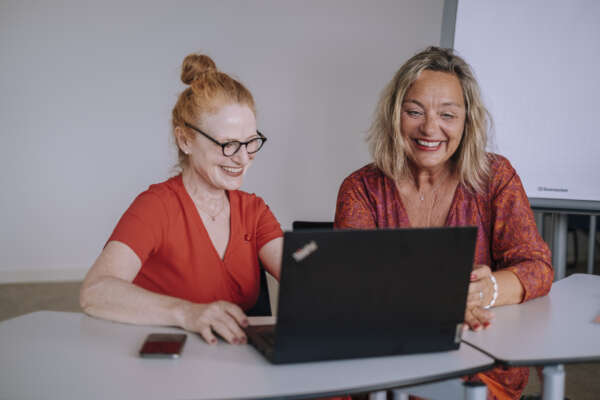 Zwei Frauen sitzen in einem Arbeitsraum an einem Tisch vor einem offenem Laptop.