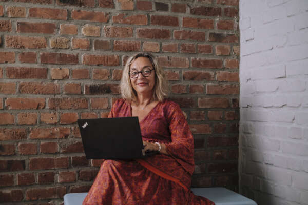 Eine Frau sitzt vor einer Backstein-Wand und hat einen offenen Laptop auf ihrem Knie liegen.