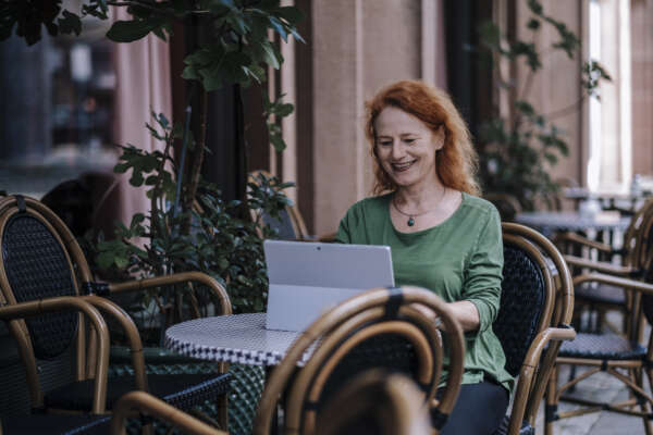 Eine lachende Frau sitzt an einem kleinen runden Tisch eines Cafés und arbeitet an einem Laptop.