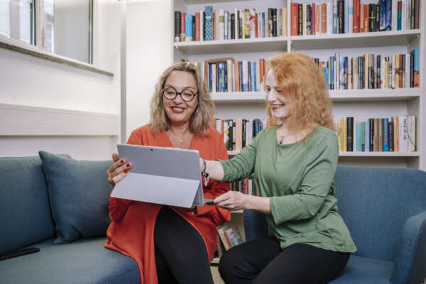 Zwei Frauen sitzen auf einer Couch vor einem Bücherregal und die linke Frau hat einen Laptop auf ihrem Schoß.