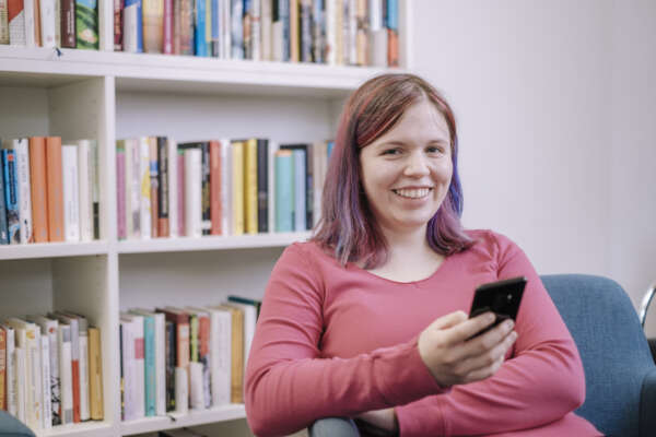 Eine Frau sitzt auf einem Sessel mit einem Handy in der Hand vor einem Bücherregal.