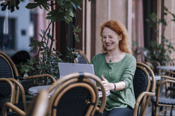 Eine lachende Frau sitzt an einem kleinen runden Tisch eines Cafés und arbeitet an einem Laptop.