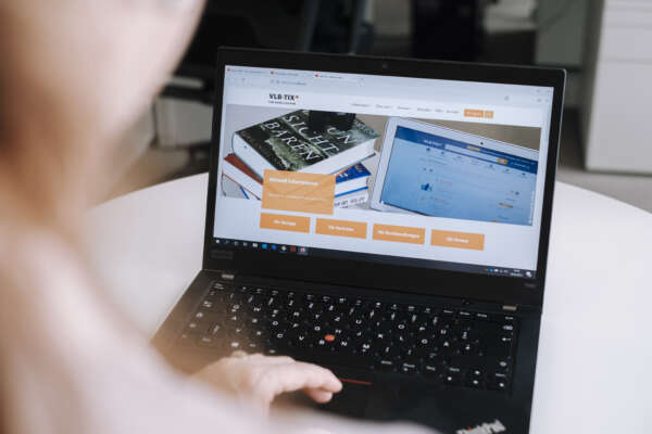 Blick über eine Schulter auf einen Laptop mit einer Hand auf der Tastatur und der Web-Oberfläche von vlbtix.de.