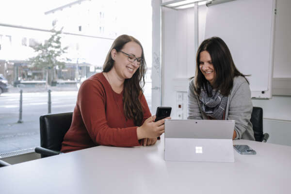 Zwei Frauen sitzen an einem runden Tisch vor einem offenem Laptop und die linke Frau hält noch ein Handy in der hand.