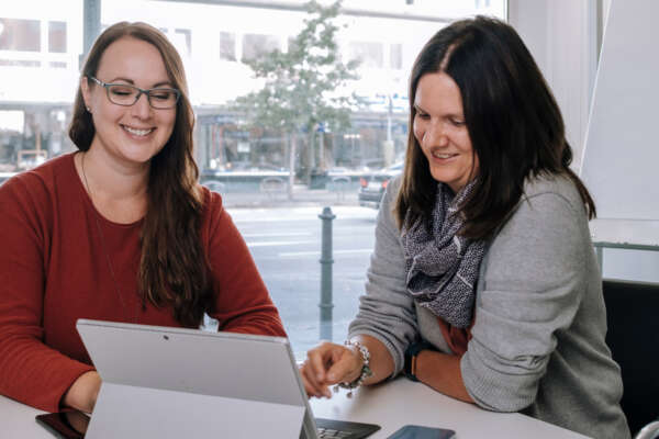 Zwei Frauen sitzen an einem Tisch vor einem offenem Laptop.