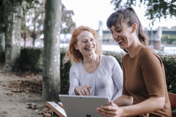 Zwei lachende Frauen sitzen auf einer Bank am Main und haben auf ihrem Schoß einen offenen Laptop liegen.