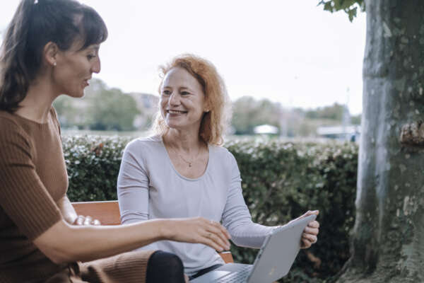 Zwei lachende Frauen sitzen auf einer Bank am Main und haben auf ihrem Schoß einen offenen Laptop liegen.